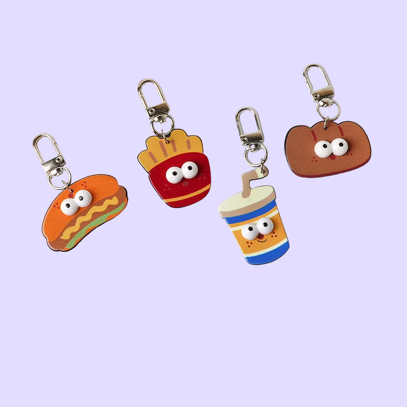 Four cute cartoon-shaped keychains with large eyes are arranged on a white surface with vintage newspaper props. The designs include a hot dog, french fries, a brown pastry, and a drink cup.