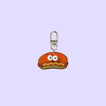 A small novelty keychain charm shaped like a smiling orange-red burger with white eyes, yellow wavy lines, and a green bottom layer, attached to a silver metal clasp.